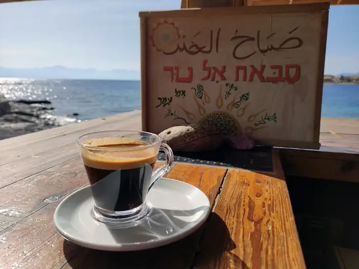 Coffee with foam in a glass on a weathered wooden table beside a hand-painted sign, with the sea beyond