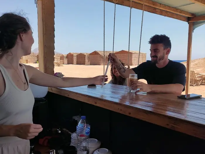 A woman at the beach bar counter being served a cold drink, thatched cabanas and sea behind